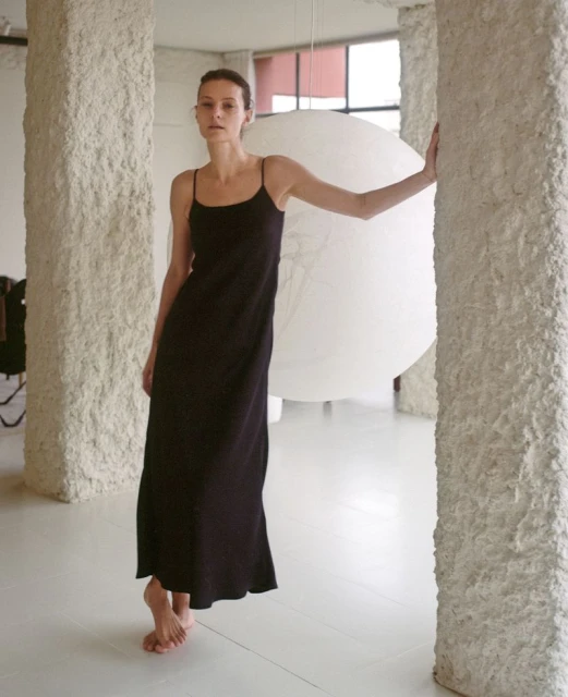 A slender woman with dark hair poses in a dark spaghetti-strap dress. She stands next to textured columns in a minimalist space.