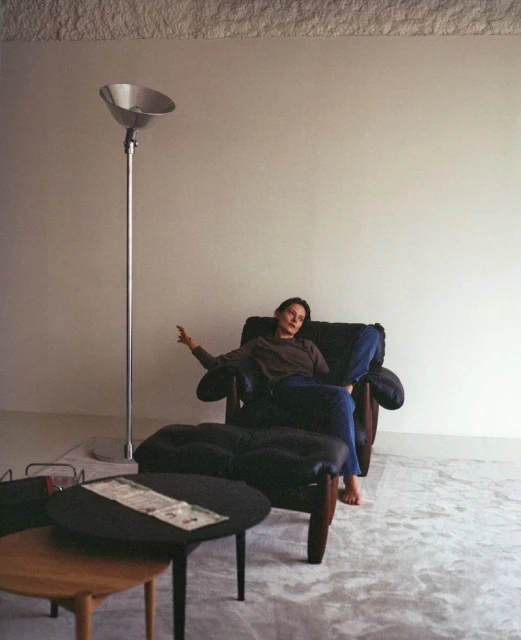 A woman sits in a black leather chair indoors with tables and a floor lamp nearby; the setting is a minimalist living room with natural light.