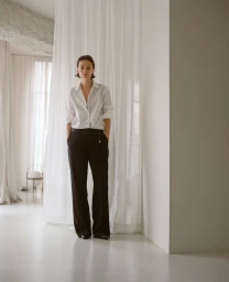 Model Tanya Ruban posing in a white button-down shirt and black wide-leg trousers in front of sheer white curtains.