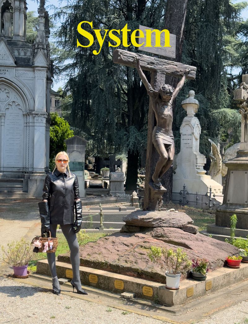A woman in a black leather jacket and gray leggings stands in a cemetery, next to a statue of Jesus on the cross, with gravestones visible.