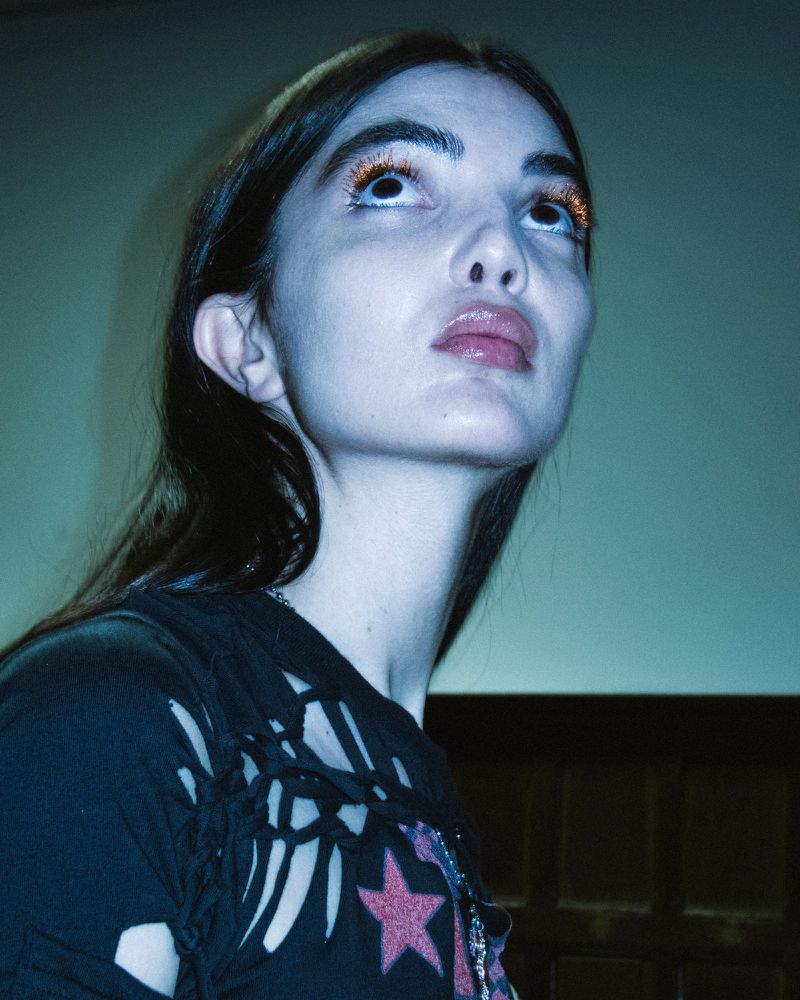 Woman with dark hair and glitter eyeshadow looks up in a medium shot in front of a blue background with a dark wooden detail.