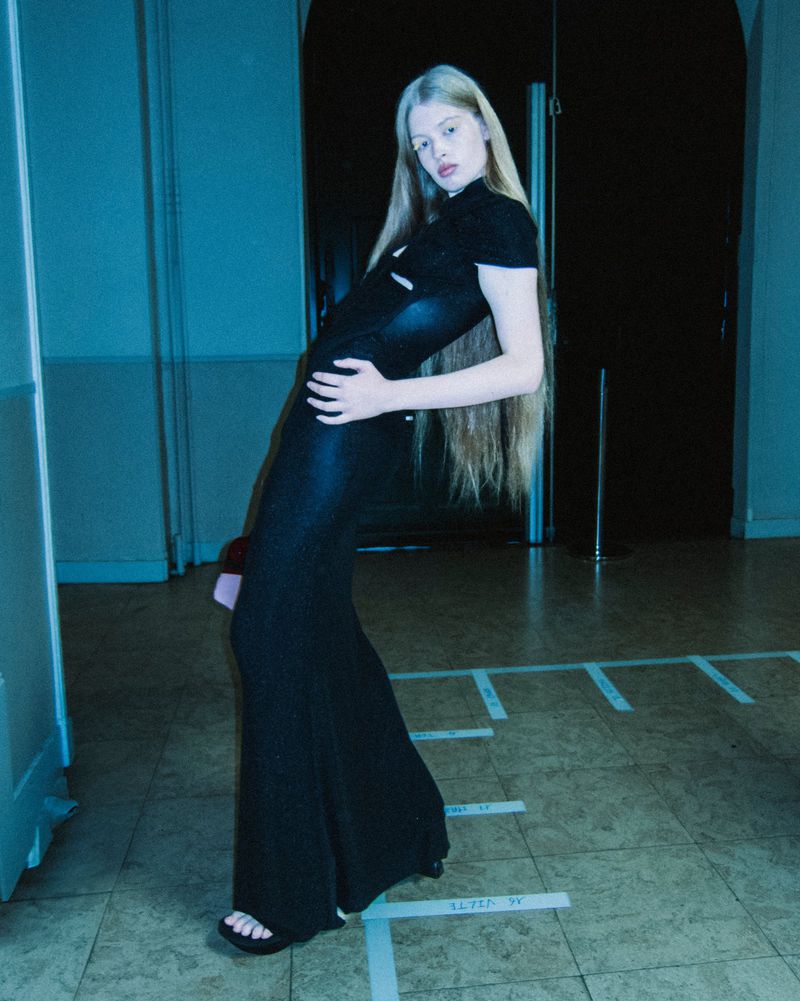 A slender woman with long blonde hair poses in a sleek black dress against a blue-toned interior backdrop with geometric floor markings.