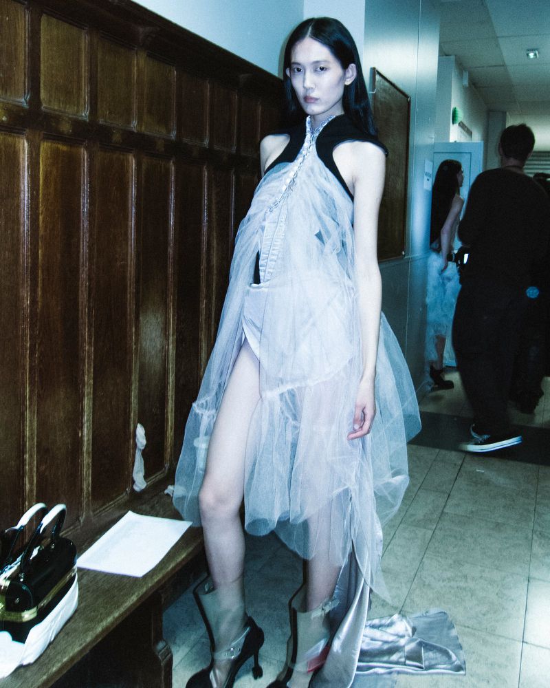 Asian fashion model in a translucent dress, standing in a hallway with wood paneling, next to a bench with shoes and other models in the background.