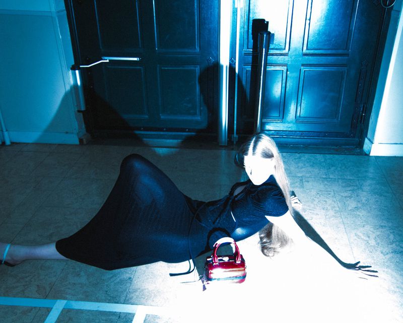 Woman lying on floor with handbag in studio. She is wearing a black dress and heels. The background features double doors.
