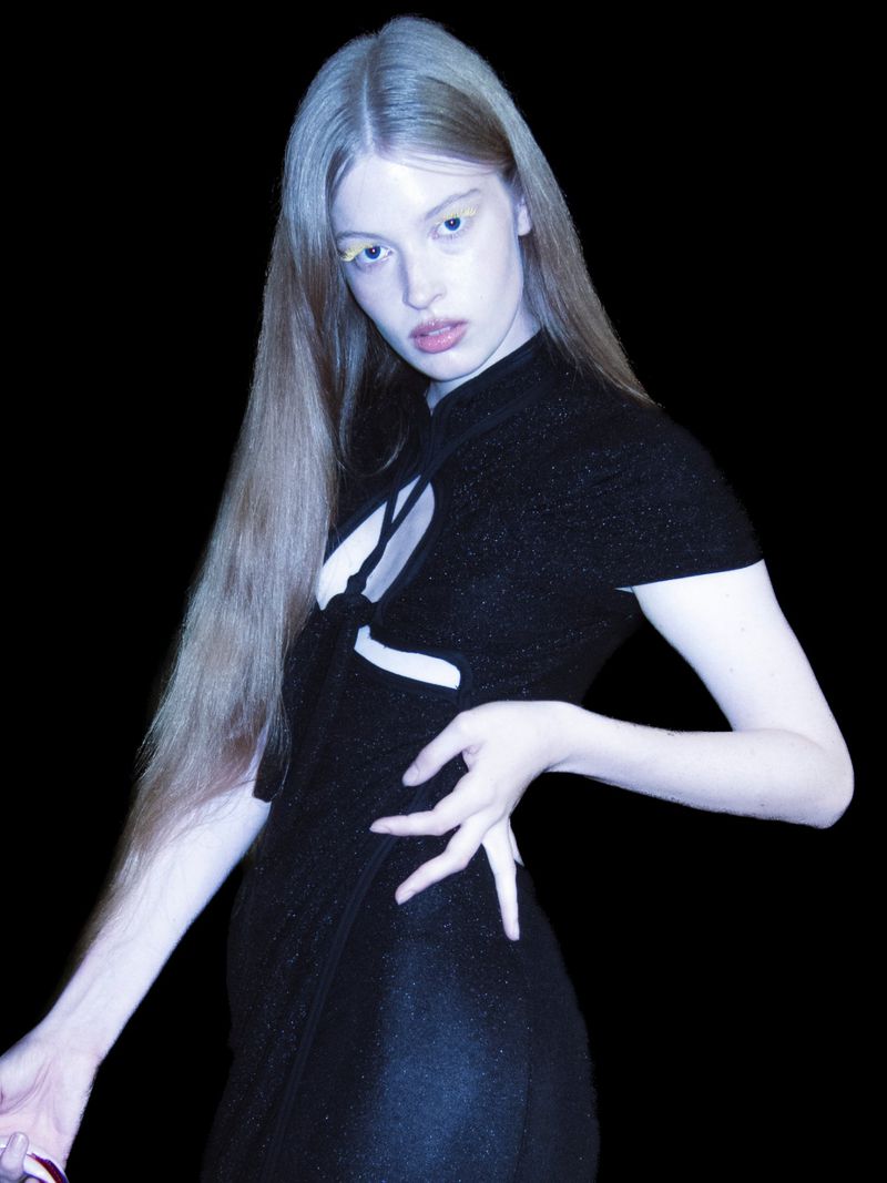 Attractive young woman with long blond hair poses in a shiny, short, black dress against a dark background, looking directly at the camera.