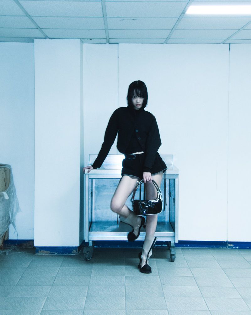A woman with short black hair wearing a dark outfit and holding a handbag poses in an indoor space with a stainless steel surface.