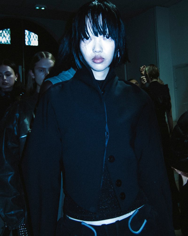 Darkly lit group of fashion models backstage wearing black clothing, medium shot focusing on a woman with dark hair and a blank expression.