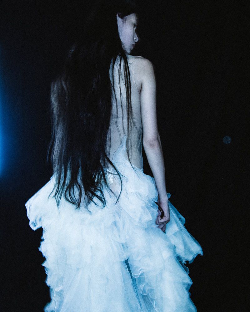 Woman in an ethereal white dress, seen from behind, with long dark hair against a dark background, fashion, beauty, and editorial.