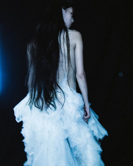 Woman in an ethereal white dress, seen from behind, with long dark hair against a dark background, fashion, beauty, and editorial.