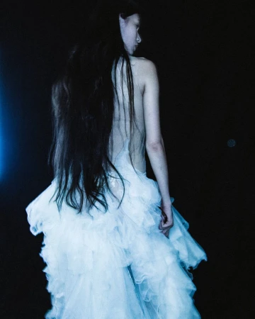 Woman in an ethereal white dress, seen from behind, with long dark hair against a dark background, fashion, beauty, and editorial.