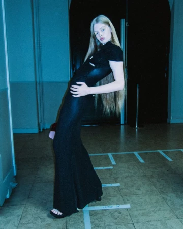 Model with long blonde hair posing in a floor-length black dress with direct flash against a dark hallway background.