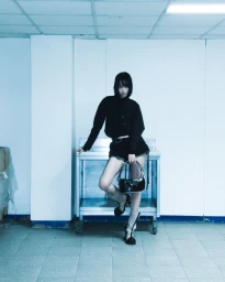 Model in black jacket and shorts posing on a metal utility cart holding a black shoulder bag against a cool-toned white wall.