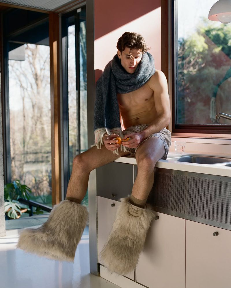 A young adult male sits on a kitchen counter wearing shorts, an unbuttoned shirt, a scarf, and furry boots while holding fruit.