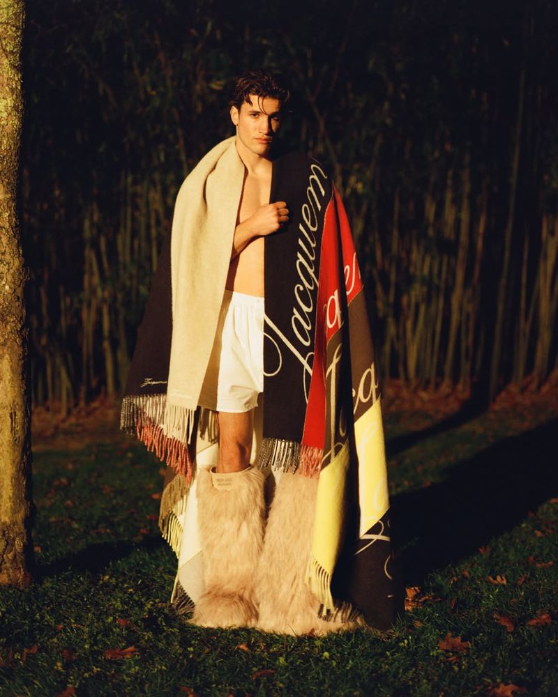 Young adult male poses outdoors with blankets and furry leg warmers. The image has a natural setting with soft lighting.