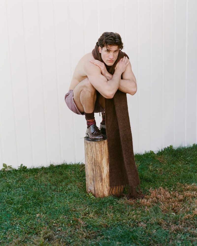 A man in shorts squats on a tree stump outdoors, wearing a scarf and shoes with socks, looking at the camera. White wall background.