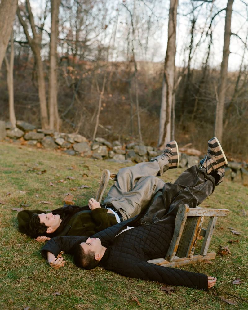 Two men lounge on a grassy field with bare trees in the background, their legs propped on an old wooden ladder, enjoying a peaceful day.