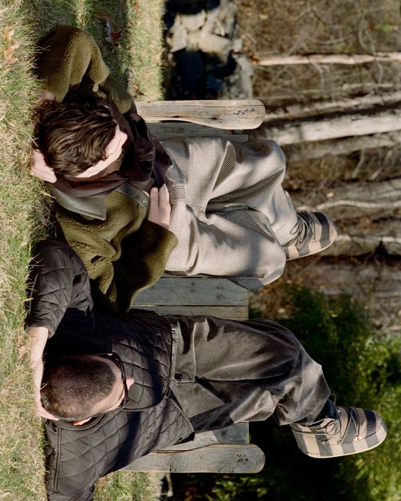 Two people relaxing outdoors on a wooden structure, one wearing a quilted jacket, the other in earth-toned layers, surrounded by grass and trees.