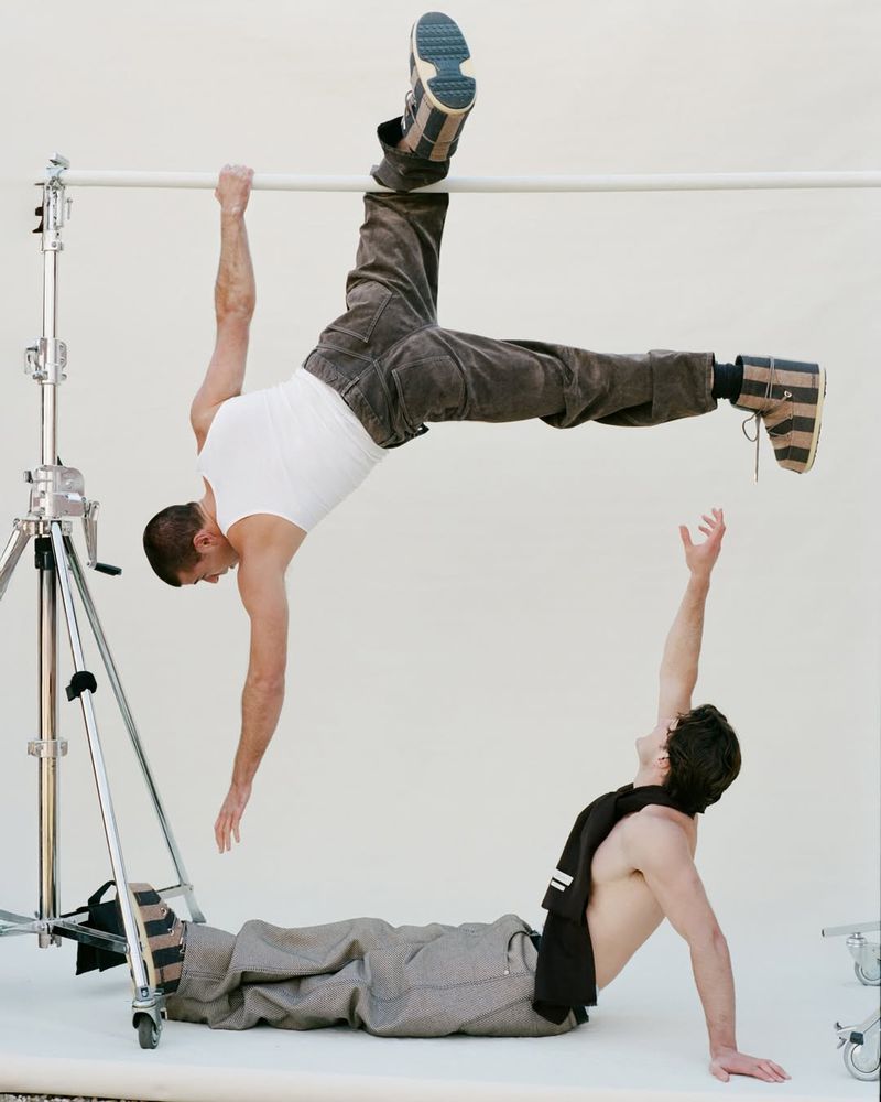 Two men are in a photo studio; one hangs from a bar, and the other supports himself with arms while laying down, showcasing casual fashion.