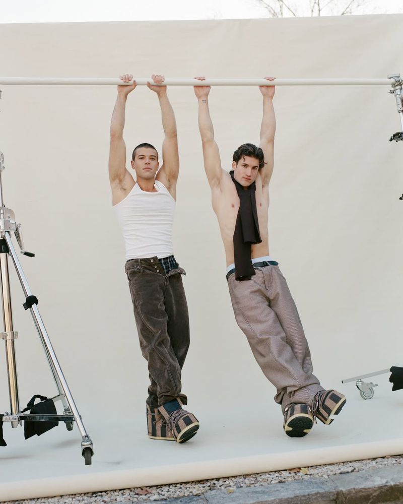 Two shirtless men in pants and platform shoes hang from a bar against a neutral backdrop in a brightly lit photo studio setting.