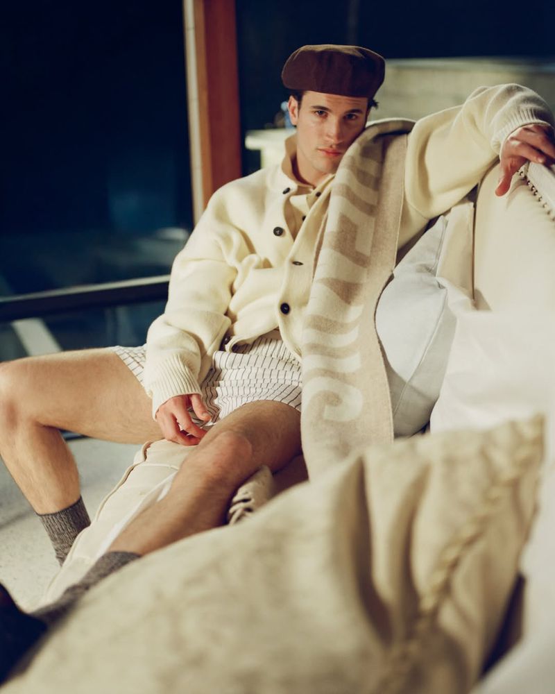 An adult male in a cardigan and hat poses while sitting on a white couch in an indoor setting with visible natural lighting.