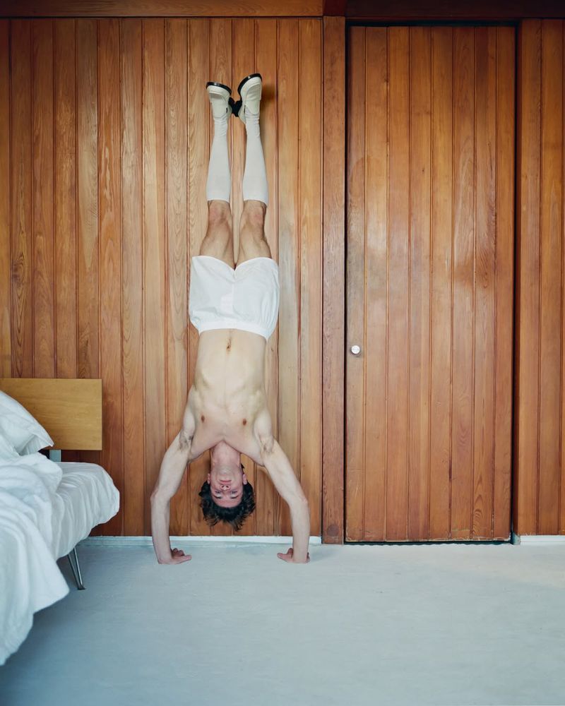 Adult male performs a handstand in a bedroom with wooden paneling and white bedding, showcasing fitness and balance in an indoor setting.