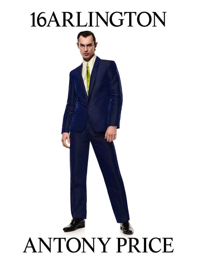 A man poses in a dark blue suit, cream shirt, and patterned tie against a white backdrop, as part of a 16ARLINGTON campaign.