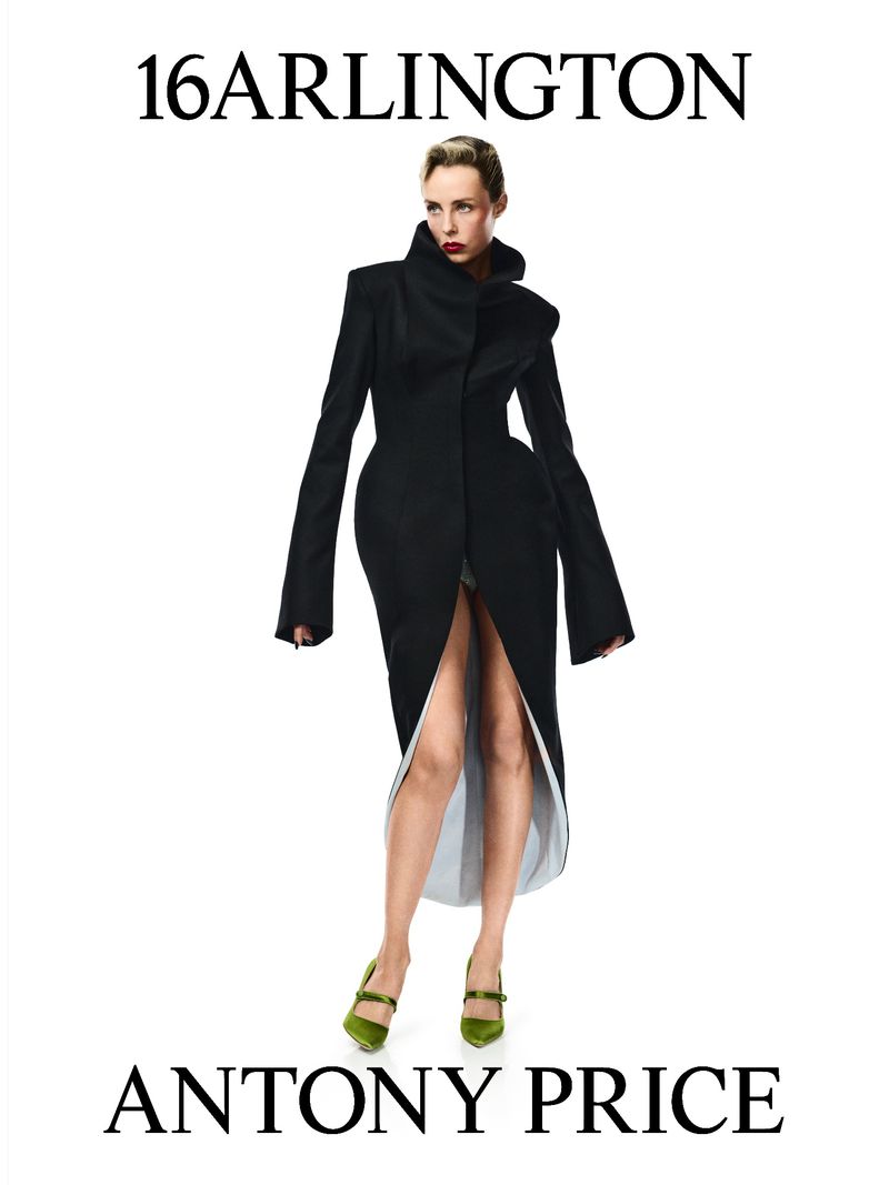 Fashion editorial shot of a woman modeling a black coat with wide sleeves, posing in front of a plain white background with visible text.