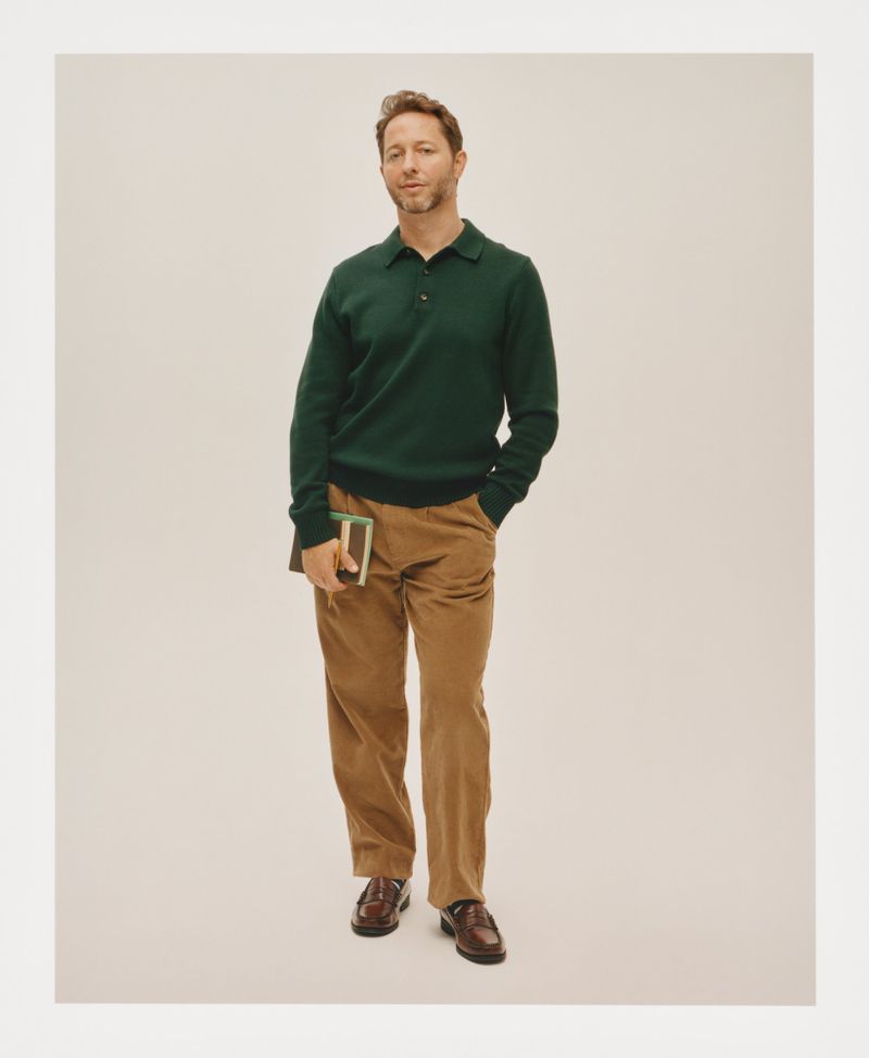 Adult male in a green polo shirt and tan pants stands holding a book in a studio with a neutral backdrop, showcasing fashion and style.