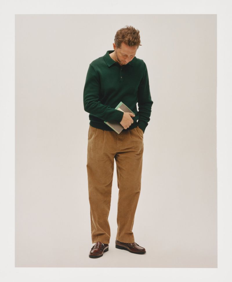 Full-length shot of an adult male in a green polo sweater and tan corduroy pants, holding a tablet against a light background.