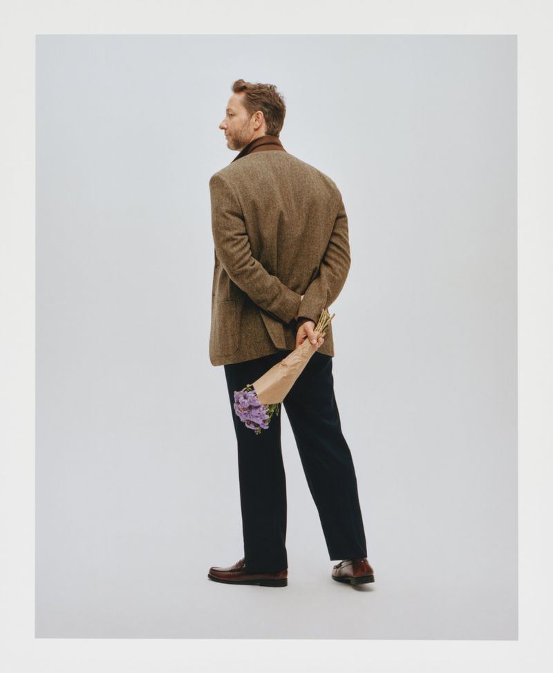 Man in tweed jacket seen from behind holds purple flowers wrapped in paper. Brown leather shoes. Neutral background.