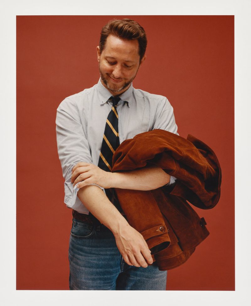 Adult male in collared shirt with tie holding a brown suede jacket against a red-brown backdrop in a studio environment. 