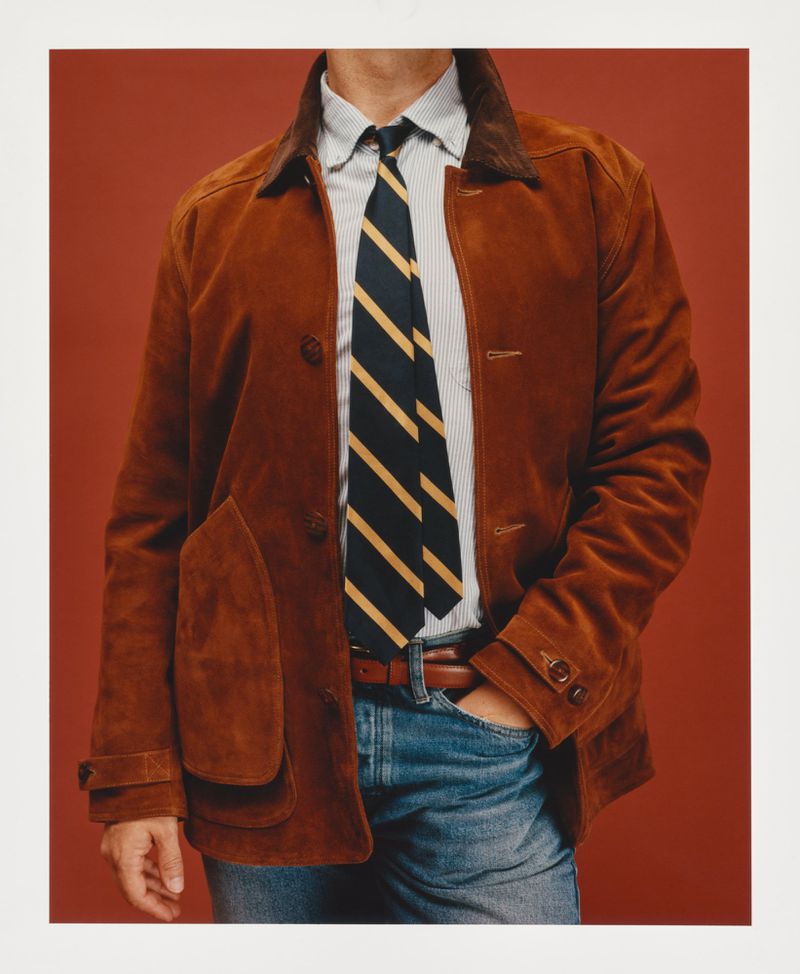 Studio shot of a man's torso wearing a brown suede jacket, striped shirt, blue and yellow striped tie, and jeans against an orange background.