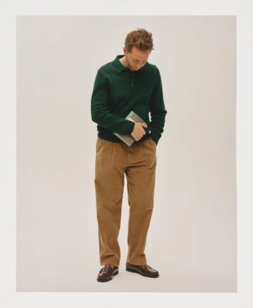Derek Blasberg in a dark green knit polo and camel pleated trousers holding a book against an off-white studio background.