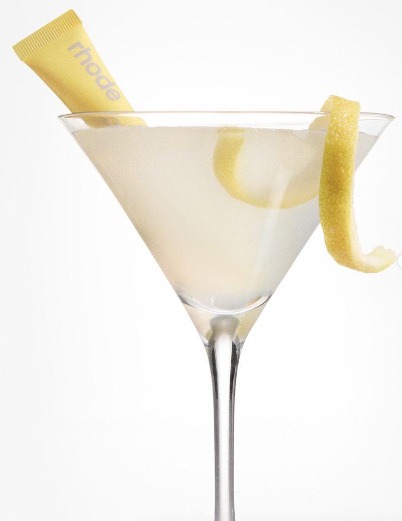 Close-up of a martini glass filled with a pale yellow liquid, garnished with a lemon twist and a yellow tube on a white background.