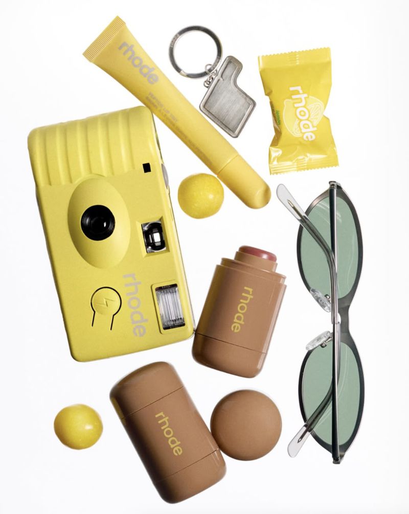 Still life arrangement of yellow camera, skincare sticks and tubes, sunglasses, and keychain, on a white background, for Rhode brand.