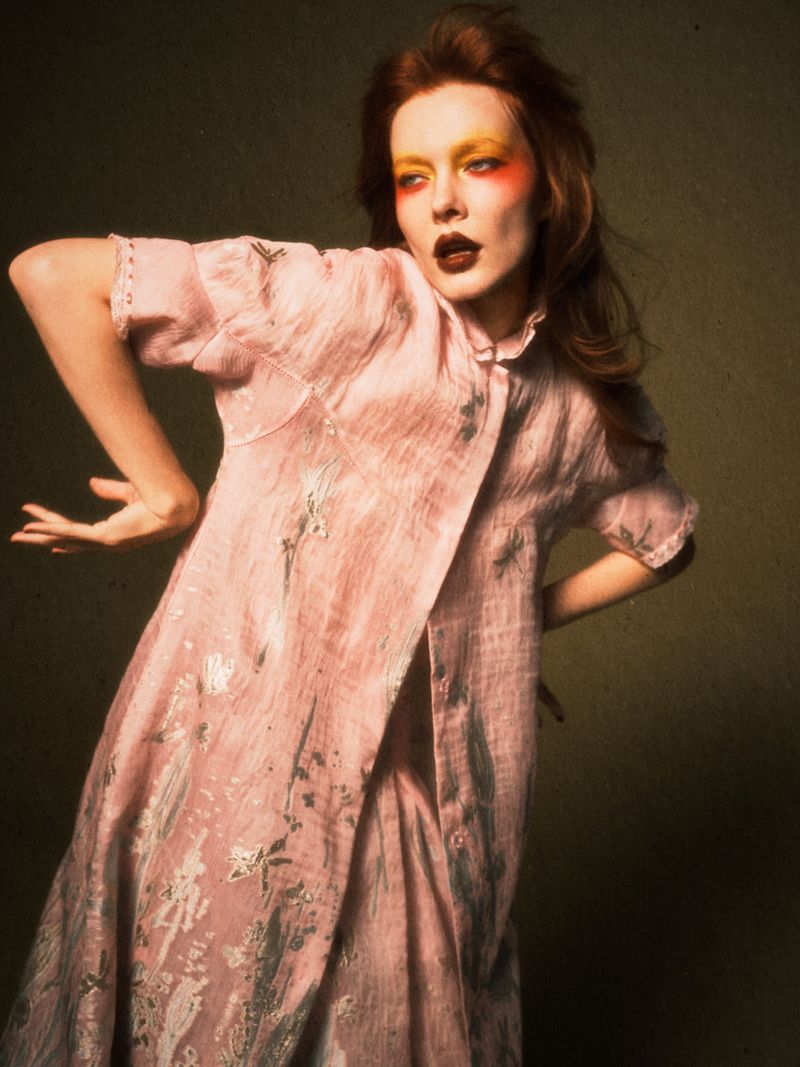 Woman in pink floral robe poses with colorful eyeshadow. Close-up portrait with red hair and dark lipstick in a photo studio setting.