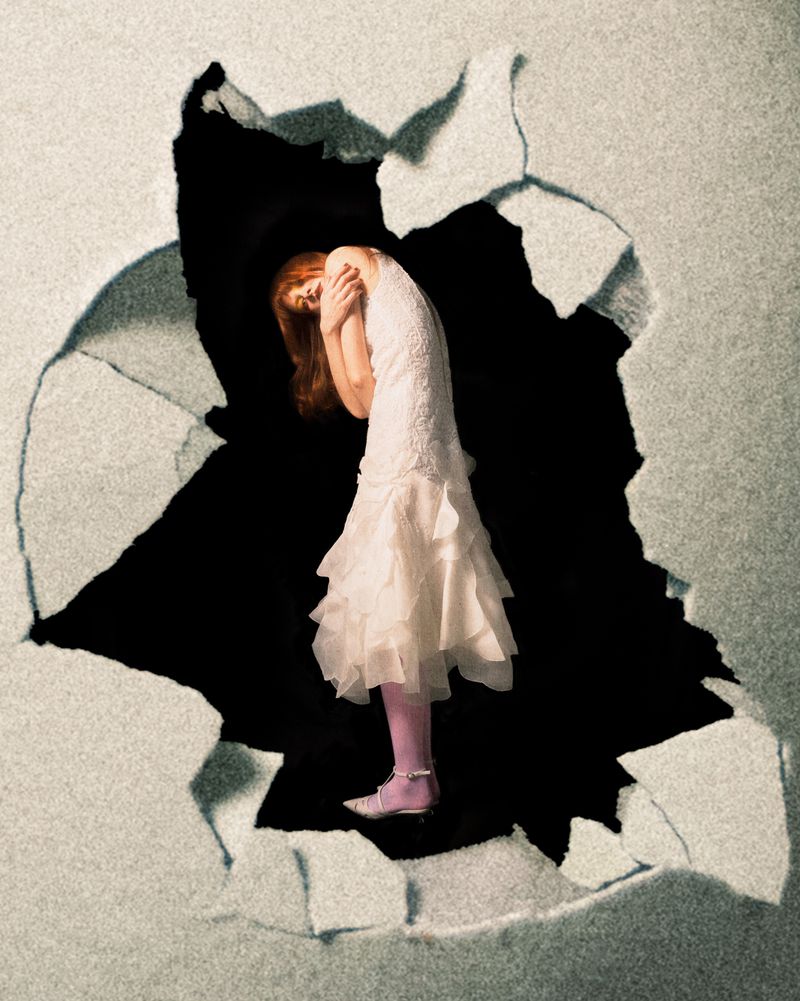 Artistic image of a red-haired woman in a white dress emerging from a cracked wall against a black background in a studio setting.