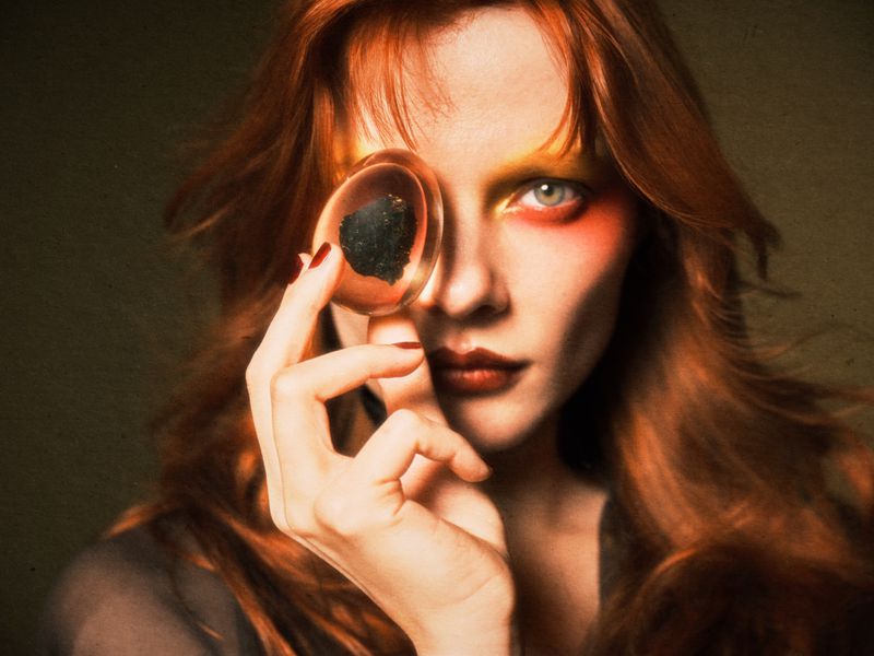 A young adult woman with red hair and vibrant makeup holds a magnifying glass to her face in a close-up beauty shot. The background is blurred.