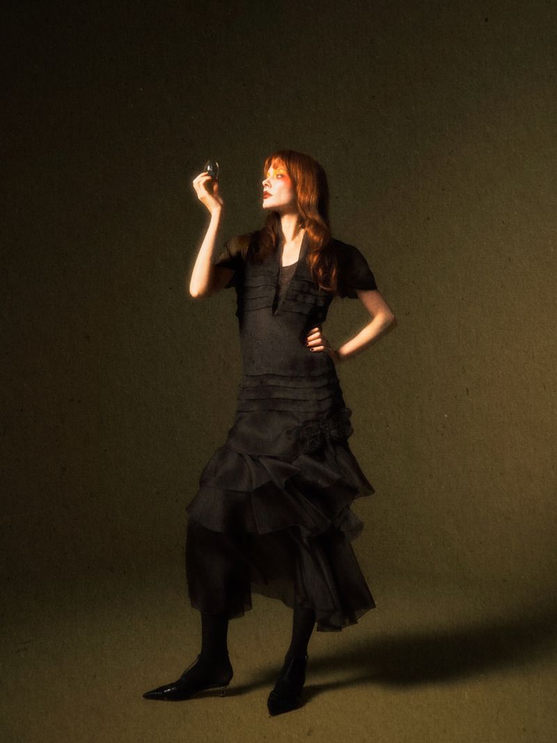 An adult woman in a ruffled black dress poses indoors, holding a small object and looking upwards in a fashion editorial style photo.