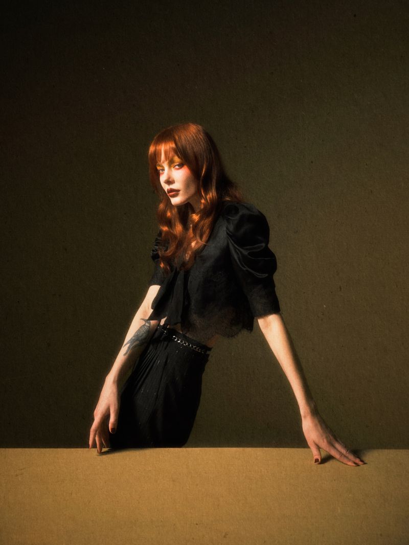 Portrait of a red-haired woman in a black blouse and pants leaning against a surface in a studio with warm, directional lighting.