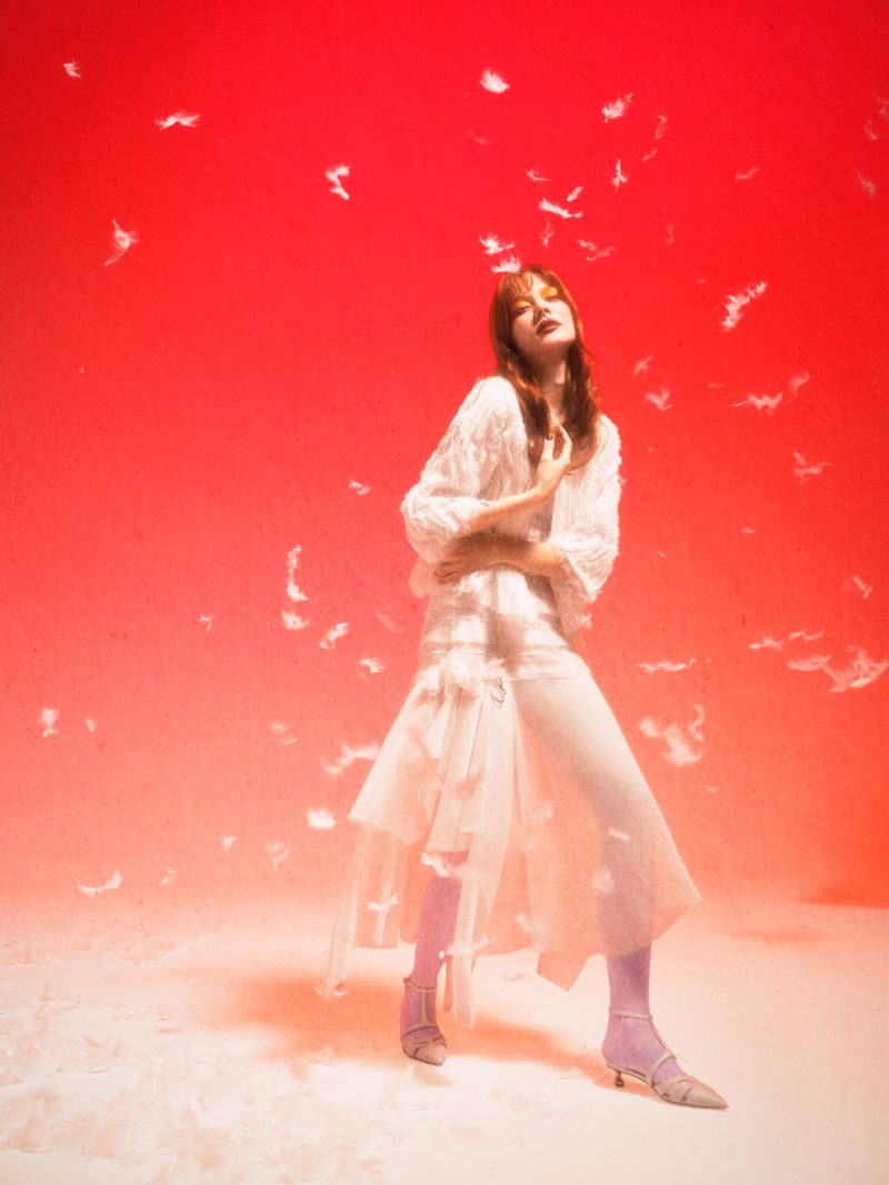 Woman in white dress poses against a red background with falling feathers. She wears kitten heels and pink tights in a fashion photography shot.