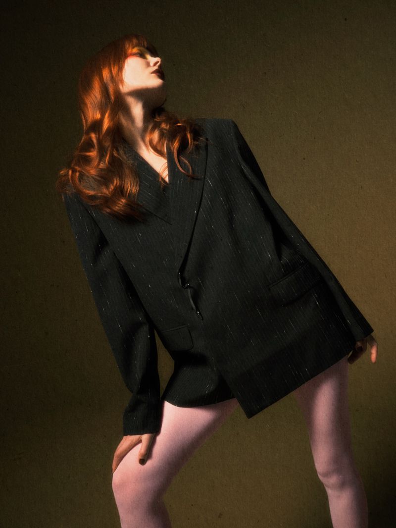 Fashion portrait of a woman with red hair wearing a black pinstripe suit jacket and pink tights against a dark olive green background.