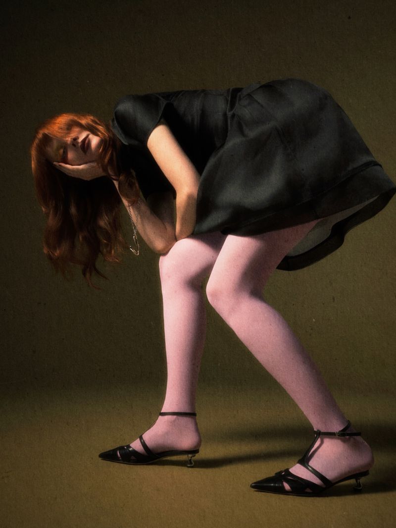 A red-haired woman wearing a black dress and pink tights poses with her hand on her face in a studio with a dark green background.
