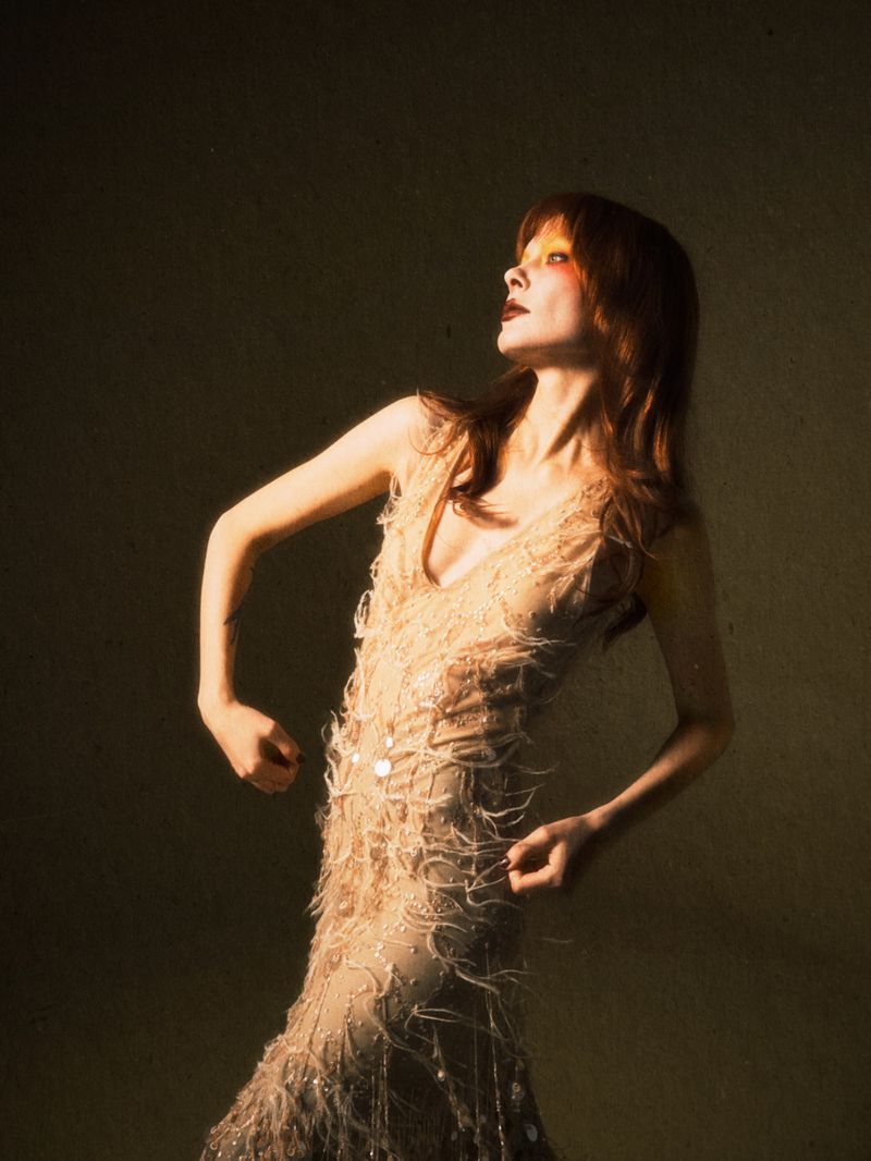 A slender woman with auburn hair and dark lipstick poses in a sequined dress, arms raised, against a muted beige studio backdrop in soft light.