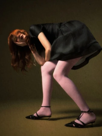 Model with long auburn hair bending forward in a black bubble mini dress, blush pink tights, and strappy black heels against an olive backdrop.