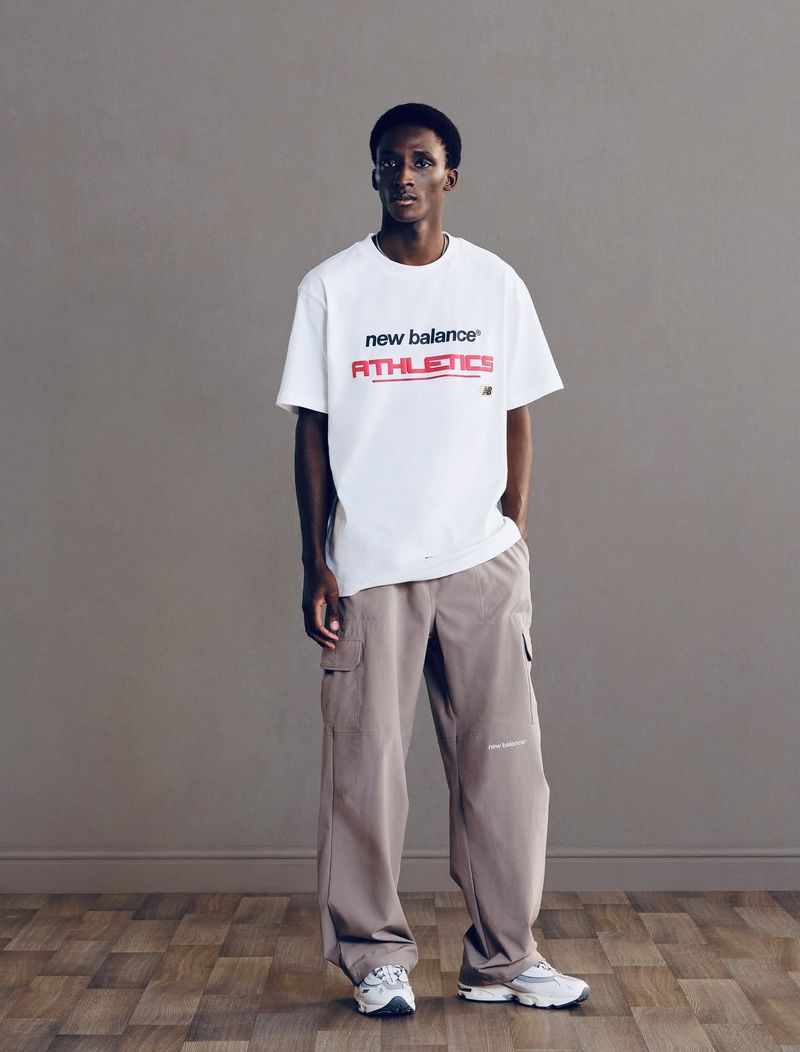 Full-length studio shot of a slender adult male wearing a white New Balance Athletics t-shirt, khaki cargo pants, and running shoes.
