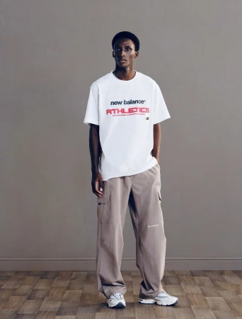 Full-length portrait of a black man wearing a white t-shirt and grey trousers standing on wooden flooring against a plain grey wall background
