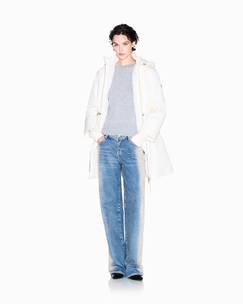 A young adult fashion model poses in a photo studio, wearing a white coat, gray sweater, and light blue jeans against a clean white backdrop.