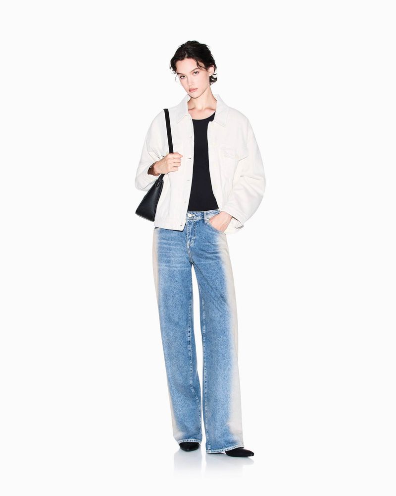 A full-length studio shot of a young adult woman in jeans, a black top, and a jacket, standing in front of a clean white backdrop.