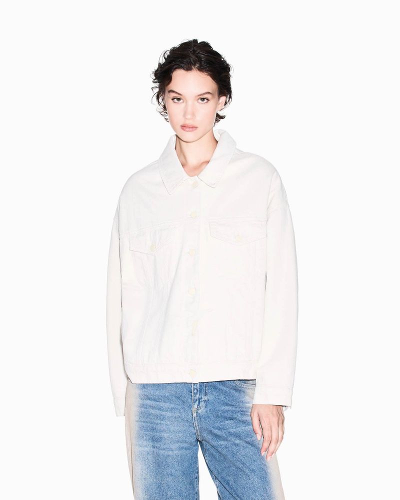 A young adult female poses in a studio with a white backdrop, wearing a light beige jacket and blue denim jeans, looking at the camera.